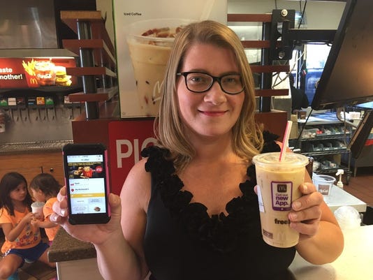 A woman in a black shirt with glasses holds an iced coffee and a cellphone up for the camera in a McDonald's. Photo Credit:  Rhonda Grundemann, The Grundemann Group